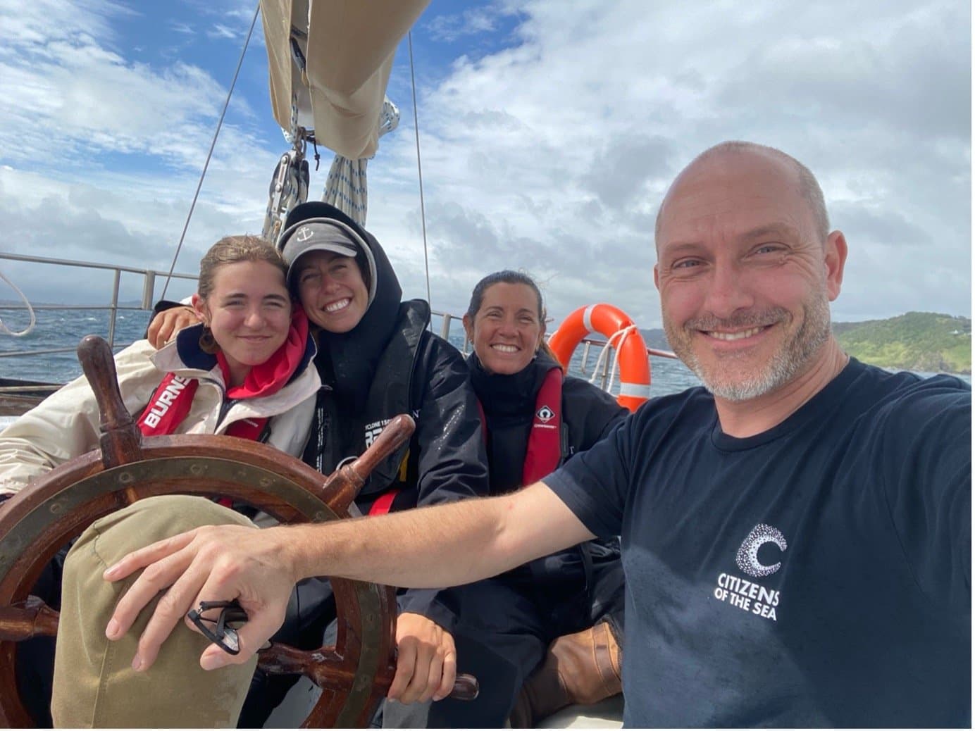 Four crew smiling at the wheel of a yacht under sail, one in a Citizens of the Sea shirt, with a coastline behind